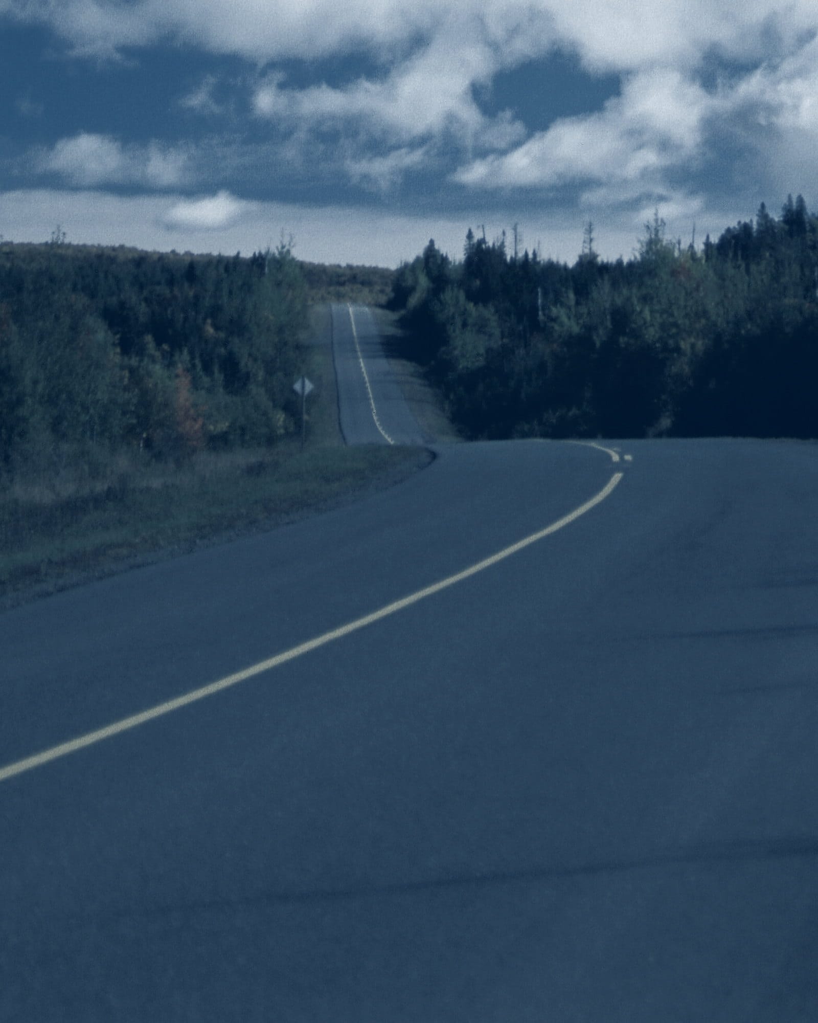An empty two-lane highway curving through New Brunswick forest under a low, cold sky